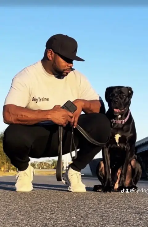 CJ kneeling beside a calm black dog on a leash outdoors